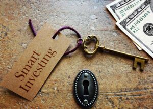 Skeleton key with a piece of paper that reads “Start Investing” attached to it by string, resting on a table next to a lock.