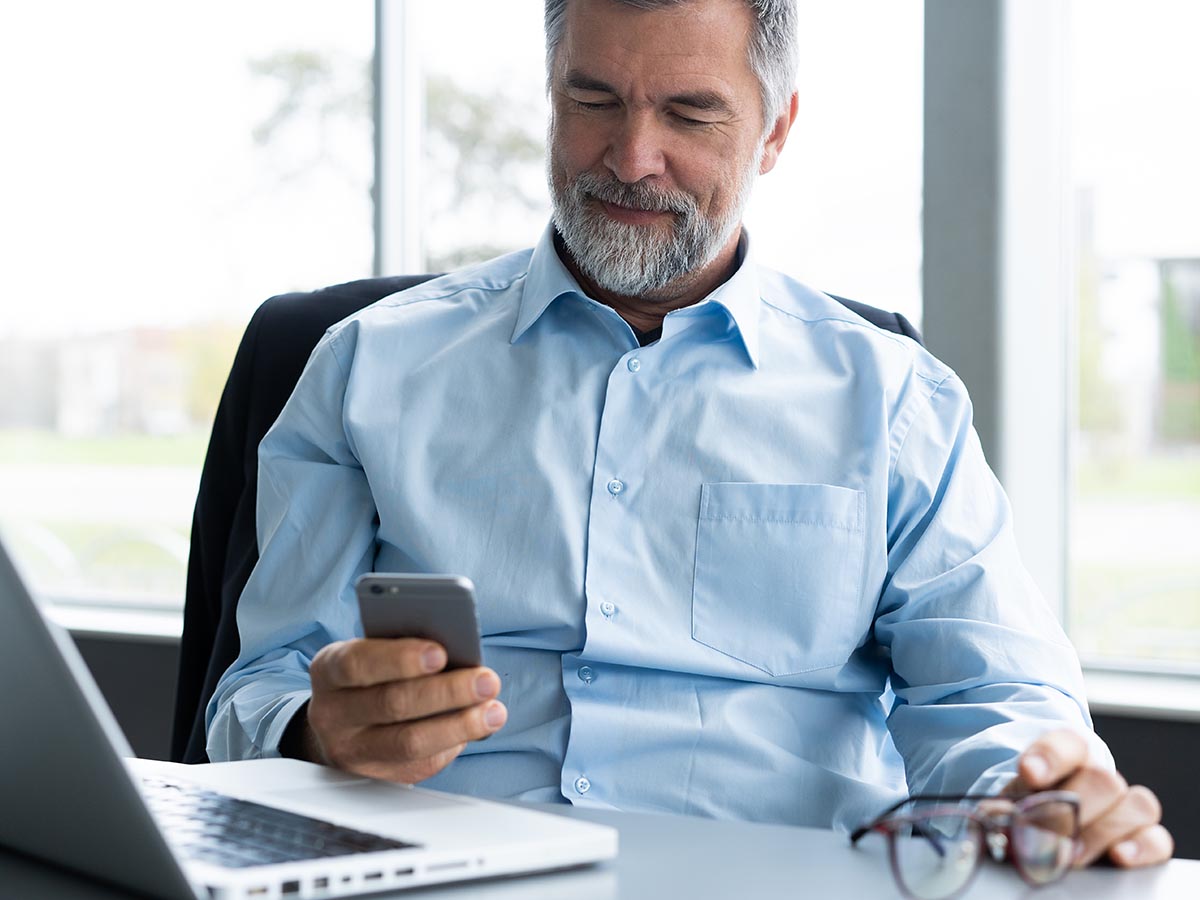 Happy senior man sitting at his desk with a laptop using his mobile phone