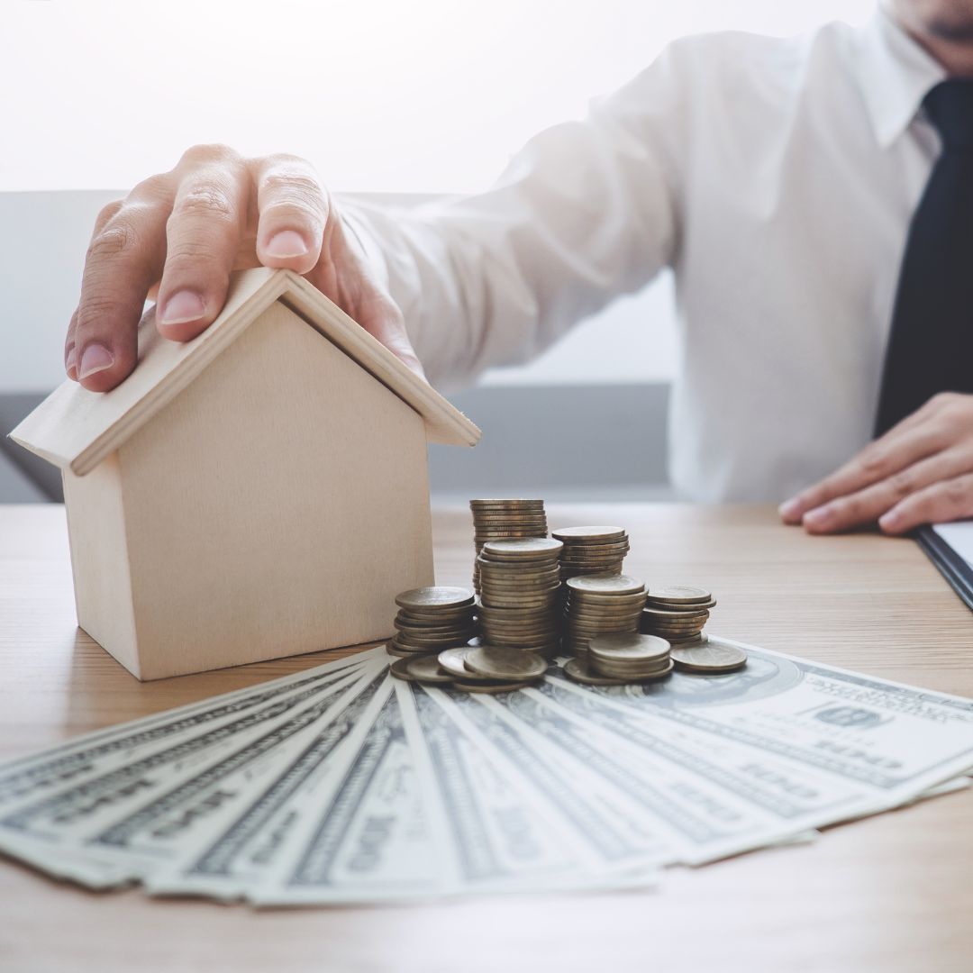a person with a small wooden house on a desk and a fan of money