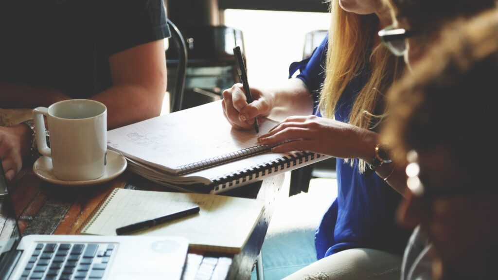 A diverse group of real estate professionals sits around a table, reviewing documents with a modern residential development in the background.