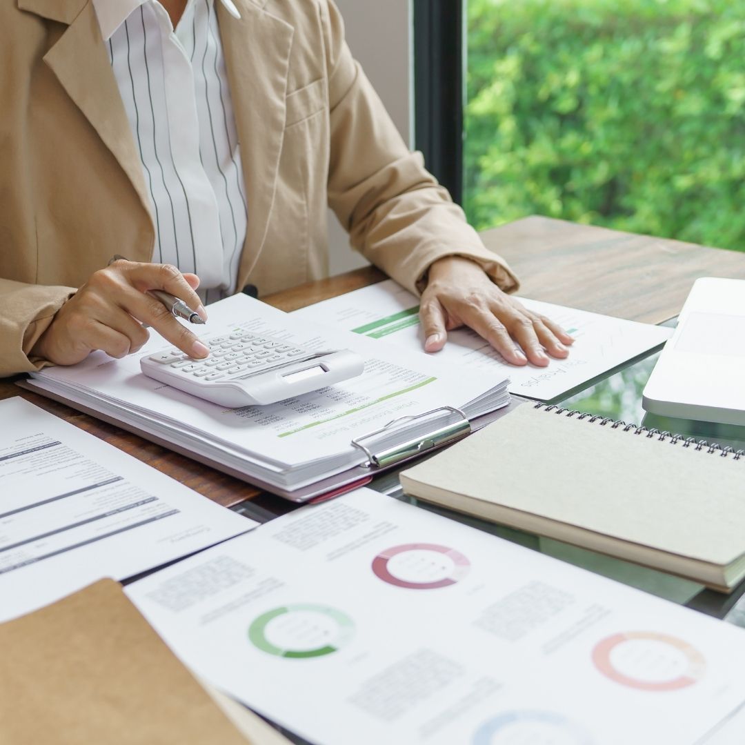 A person in a tan blazer uses a calculator while working at a desk with financial papers and charts.