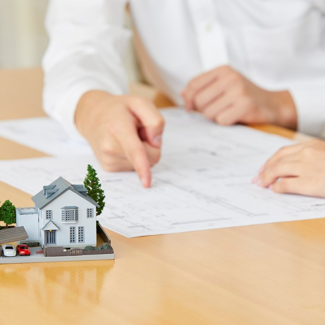 A person in a white shirt points to a floor plan on a tablet next to a detailed house model and miniature cars.