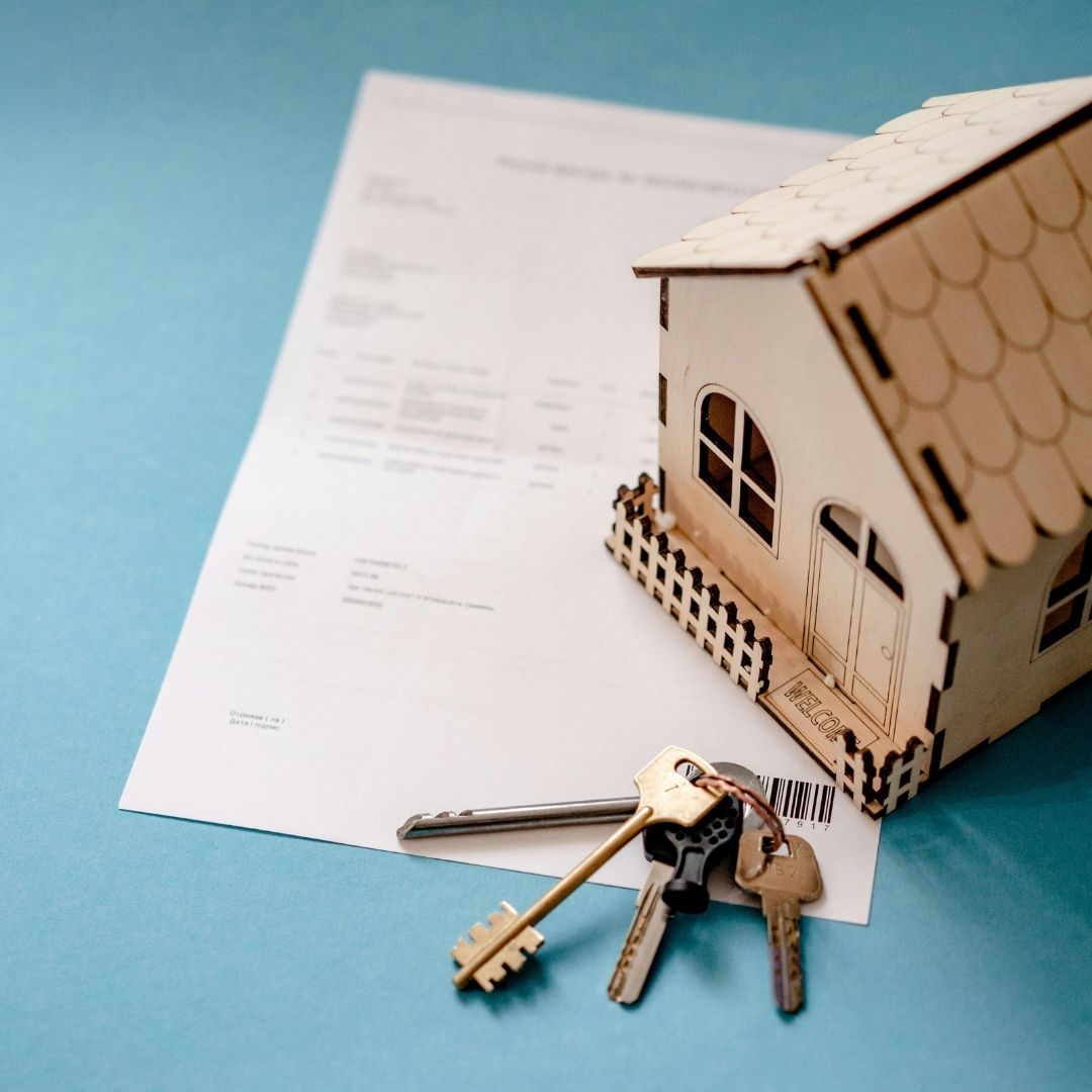 A top-down view of a small wooden house model and a set of keys on a white contract paper against a blue background.