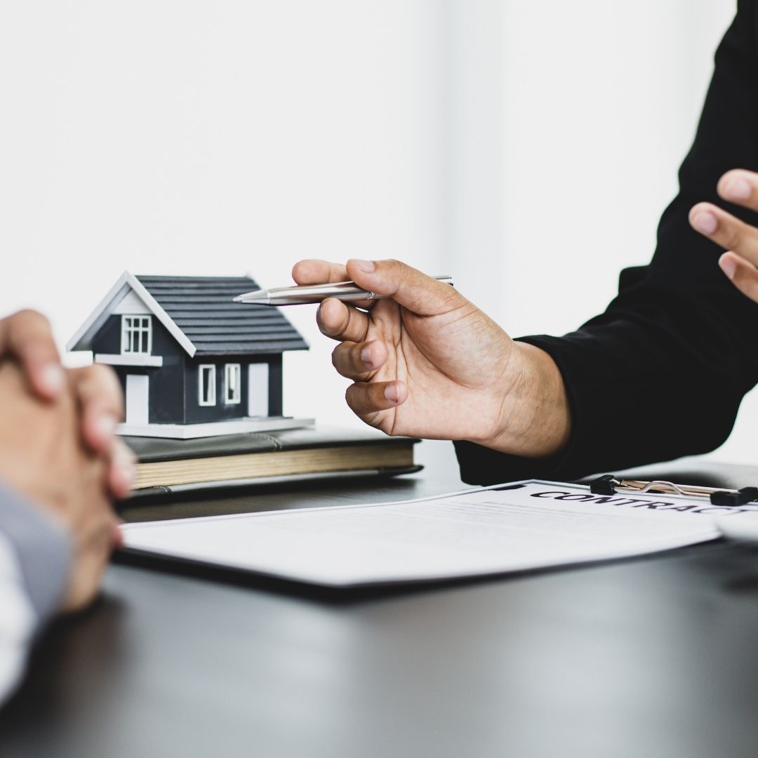 Two people sit at a desk with their hands near a real estate contract; one person points a pen toward the document while a miniature house model sits nearby.